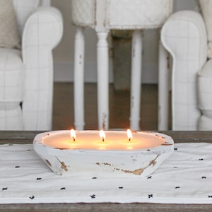 May include: A white, oval-shaped wooden dough bowl candle featuring three lit wicks. The exterior of the bowl has a distressed, weathered finish with patches of exposed brown wood showing through the white paint, containing smooth, cream-colored wax.