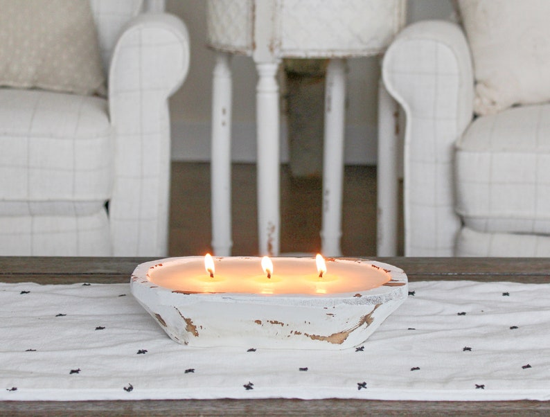 May include: A white, oval-shaped wooden dough bowl candle featuring three lit wicks. The exterior of the bowl has a distressed, weathered finish with patches of exposed brown wood showing through the white paint, containing smooth, cream-colored wax.
