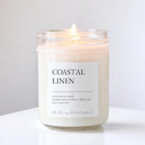 May include: A lit soy candle in a clear glass jar with a white label reading Coastal Linen, 100% pure soy candle, handpoured at our farm in Bixby, OK, 40 hr. burn time, and The Dancing Wick Candle Co. The wax is off-white and the flame is visible.