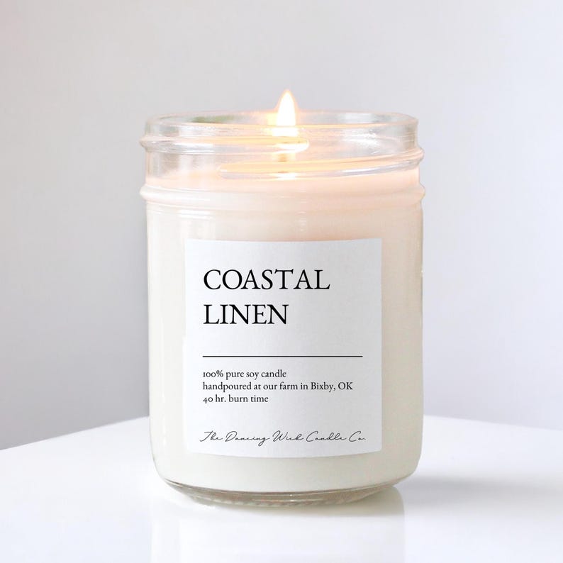 May include: A lit soy candle in a clear glass jar with a white label reading Coastal Linen, 100% pure soy candle, handpoured at our farm in Bixby, OK, 40 hr. burn time, and The Dancing Wick Candle Co. The wax is off-white and the flame is visible.