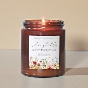 May include: A lit, brown glass jar candle with a white label. The label reads "be still & know that I am God" with floral accents. The candle is 8 oz and hand-poured in Bixby, OK. The Dancing Wick Candle Co.
