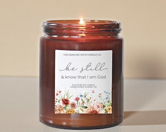 May include: A lit, brown glass jar candle with a white label. The label reads "be still & know that I am God" with floral accents. The candle is 8 oz and hand-poured in Bixby, OK. The Dancing Wick Candle Co.