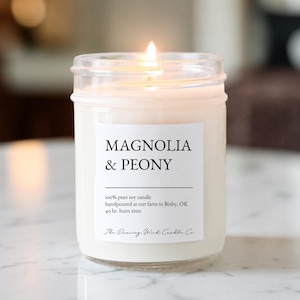May include: A lit candle in a clear glass jar with a white label reading Magnolia & Peony, 100% pure soy candle, handpoured at our farm in Bixby, OK, 40 hr. burn time, and The Dancing Wick Candle Co. The wax is off-white and the wick is burning.