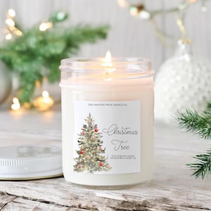 K&ouml;nnte beinhalten: Eine klare Glaskerze in einem Einmachglas mit einem wei&szlig;en Etikett, auf dem "The Dancing Wick Candle Co." und "Christmas Tree" steht. Das Etikett zeigt auch eine Aquarell-Illustration eines Weihnachtsbaums mit roten Kugeln und Lichtern. Die Kerze besteht aus 8 oz reinem Soja und wird in Boise, ID, handgefertigt.