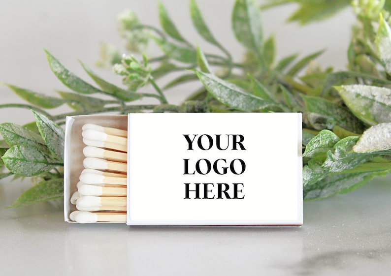 Pu&ograve; includere: Una scatola di fiammiferi bianca con un'etichetta bianca che dice "YOUR LOGO HERE" e una fila di fiammiferi di legno con punte bianche. La scatola di fiammiferi &egrave; su una superficie bianca con del fogliame verde dietro.