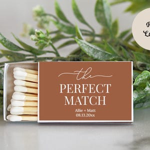 Peut inclure: Une bo&icirc;te d'allumettes rectangulaire avec une &eacute;tiquette couleur rouille portant le texte blanc "the PERFECT MATCH", les pr&eacute;noms "Allie + Matt" et la date "08.13.20xx". La bo&icirc;te est entrouverte, r&eacute;v&eacute;lant des allumettes en bois &agrave; bouts blancs.