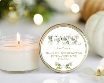 May include: A lit candle in a clear glass jar next to a round, gold-rimmed lid. The lid features the words "FALL in love" with floral accents and text that reads "THANK YOU FOR SHOWERING KATRINA WITH LOVE 10.24.20xx".