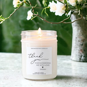 Puede incluir: Una vela de soja blanca en un tarro de vidrio transparente con una etiqueta que dice "The Dancing Wick Candle Co." y "Gracias por ducharme con amor Sarah".