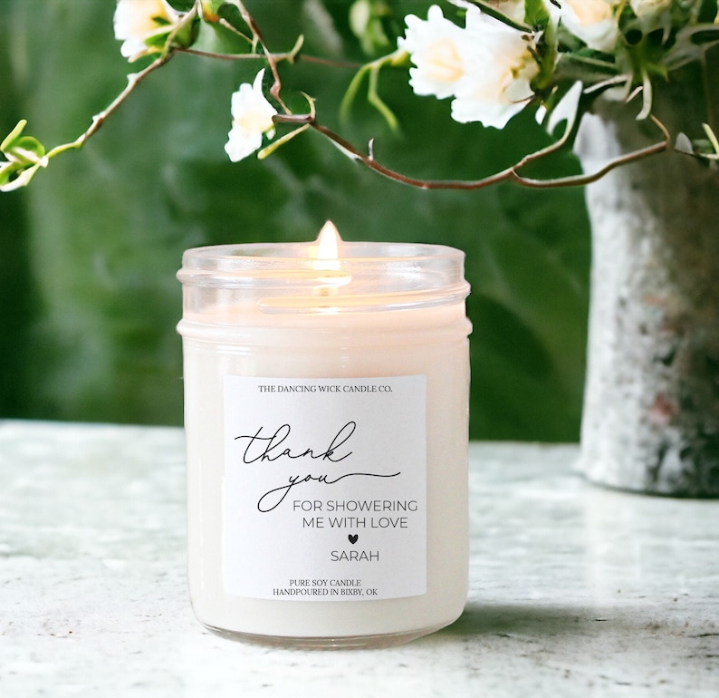 Puede incluir: Una vela de soja blanca en un tarro de vidrio transparente con una etiqueta que dice "The Dancing Wick Candle Co." y "Gracias por ducharme con amor Sarah".
