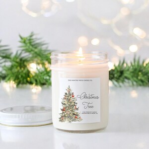 K&ouml;nnte beinhalten: Eine wei&szlig;e Kerze in einem durchsichtigen Glasgef&auml;&szlig; mit einem wei&szlig;en Etikett, auf dem "The Dancing Wick Candle Co." und "Christmas Tree" steht. Das Etikett zeigt au&szlig;erdem eine Aquarell-Illustration eines Weihnachtsbaums mit roten Kugeln.