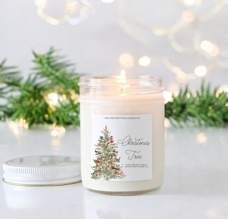 K&ouml;nnte beinhalten: Eine wei&szlig;e Kerze in einem durchsichtigen Glasgef&auml;&szlig; mit einem wei&szlig;en Etikett, auf dem "The Dancing Wick Candle Co." und "Christmas Tree" steht. Das Etikett zeigt au&szlig;erdem eine Aquarell-Illustration eines Weihnachtsbaums mit roten Kugeln.