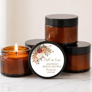 May include: A group of amber glass jars with black lids, one of which is lit and contains a candle. A circular white label on the front features a floral illustration in autumn colors and the text: Fall in Love, AMANDA'S BRIDAL SHOWER, Thanks for coming!