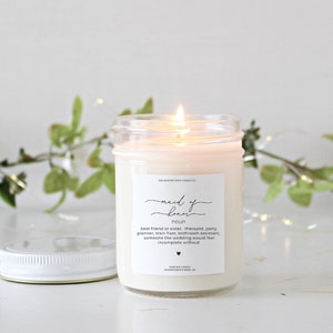 May include: A clear glass jar candle with a white label that reads "maid of honor" and a definition of the term. The candle is lit and the flame is visible.