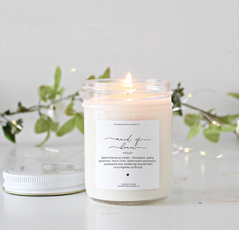 May include: A clear glass jar candle with a white label that reads "maid of honor" and a definition of the term. The candle is lit and the flame is visible.