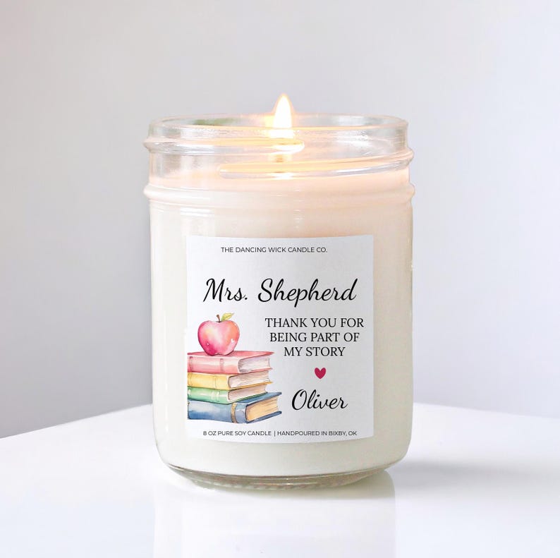 May include: A lit, white soy candle in a glass jar. The label reads "Mrs. Shepherd, THANK YOU FOR BEING PART OF MY STORY, Oliver" with an apple and books illustration. The candle is hand-poured in Bixby, OK.