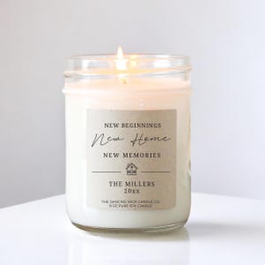 May include: A lit white soy candle in a clear glass jar. The candle has a label with the words "New Beginnings, New Home, New Memories" and "The Millers 20xx". The text "The Dancing Wick Candle Co. 8 oz Pure Soy Candle" is also on the label.