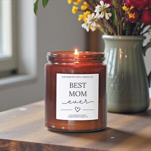 以下が含まれることがあります： 「BEST MOM ever」の文字とハートマークが入った白いラベルが付いた、火の灯った茶色のガラス瓶キャンドル。キャンドルは255gで、花の入った緑色の花瓶の隣にあります。