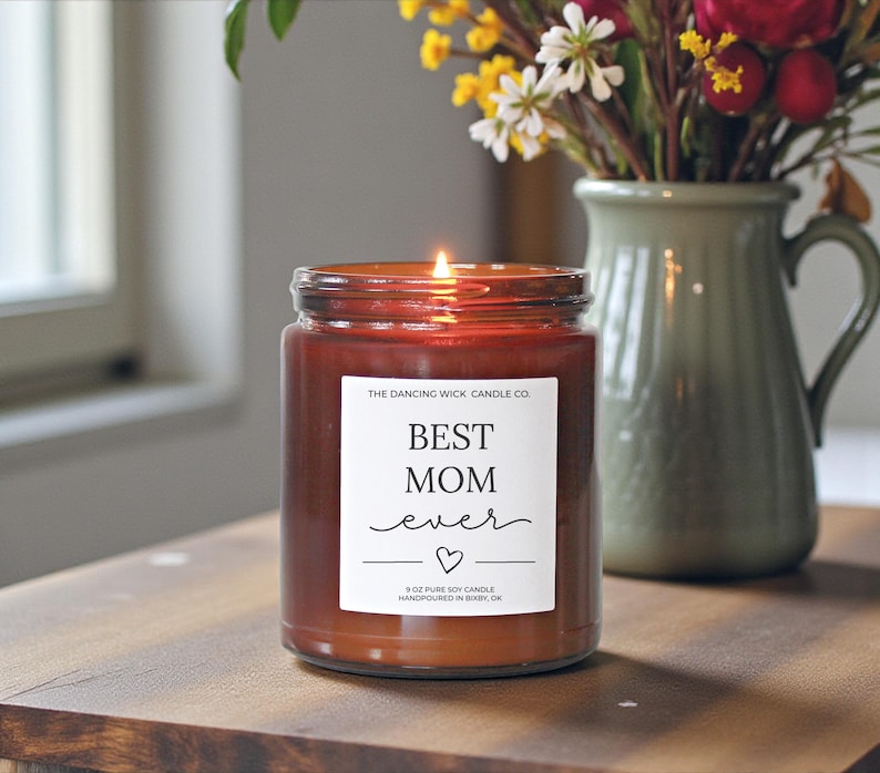 以下が含まれることがあります： 「BEST MOM ever」の文字とハートマークが入った白いラベルが付いた、火の灯った茶色のガラス瓶キャンドル。キャンドルは255gで、花の入った緑色の花瓶の隣にあります。