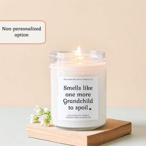 Puede incluir: Una vela de soja blanca encendida en un tarro de cristal con el texto "Smells like one more Grandchild to spoil." La vela est&aacute; sobre un peque&ntilde;o bloque de madera con flores blancas. La vela es de 227 gramos.
