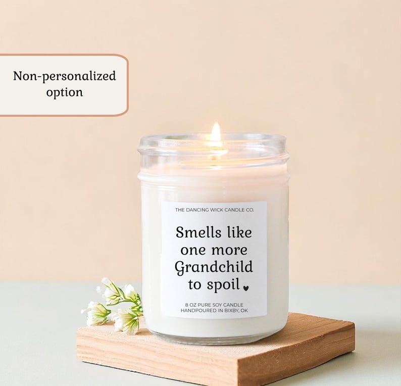 Puede incluir: Una vela de soja blanca encendida en un tarro de cristal con el texto "Smells like one more Grandchild to spoil." La vela est&aacute; sobre un peque&ntilde;o bloque de madera con flores blancas. La vela es de 227 gramos.