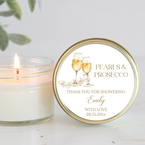 May include: A gold candle jar with a white label that says "Pearls & Prosecco" and "Thank you for showering Emily with love 08.13.20xx". The label also features a watercolor illustration of two champagne flutes.