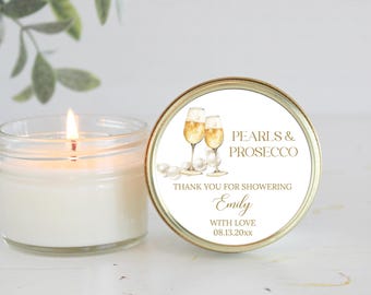 May include: A gold candle jar with a white label that says "Pearls & Prosecco" and "Thank you for showering Emily with love 08.13.20xx". The label also features a watercolor illustration of two champagne flutes.
