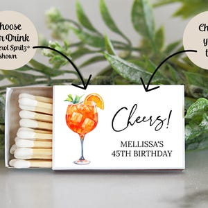 Puede incluir: Una caja de cerillas blanca con una etiqueta blanca. La etiqueta tiene una ilustraci&oacute;n de acuarela de un c&oacute;ctel Aperol Spritz y el texto "Cheers! Melissa's 45th Birthday".