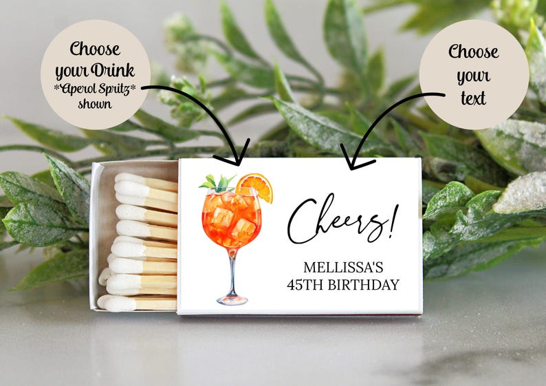 Puede incluir: Una caja de cerillas blanca con una etiqueta blanca. La etiqueta tiene una ilustraci&oacute;n de acuarela de un c&oacute;ctel Aperol Spritz y el texto "Cheers! Melissa's 45th Birthday".