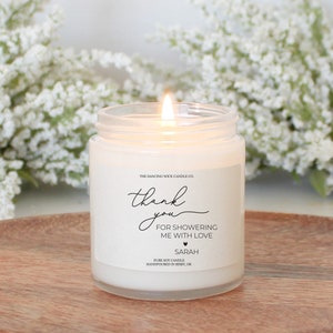 Puede incluir: Vela de soja blanca con una etiqueta que dice "The Dancing Wick Candle Co. thank you FOR SHOWERING ME WITH LOVE SARAH PURE SOY CANDLE HANDPOURED IN BIXBY, OK."