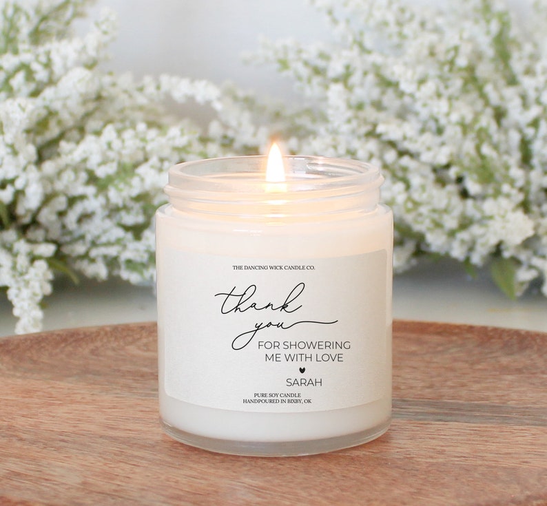 Puede incluir: Vela de soja blanca con una etiqueta que dice "The Dancing Wick Candle Co. thank you FOR SHOWERING ME WITH LOVE SARAH PURE SOY CANDLE HANDPOURED IN BIXBY, OK."