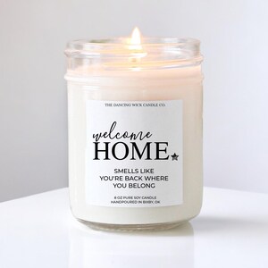 May include: A white soy candle in a glass jar with a label that reads "welcome HOME SMELLS LIKE YOU'RE BACK WHERE YOU BELONG 8 OZ PURE SOY CANDLE HANDPOURED IN BIXBY, OK"