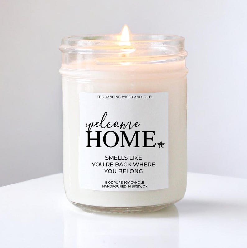 May include: A white soy candle in a glass jar with a label that reads "welcome HOME SMELLS LIKE YOU'RE BACK WHERE YOU BELONG 8 OZ PURE SOY CANDLE HANDPOURED IN BIXBY, OK"