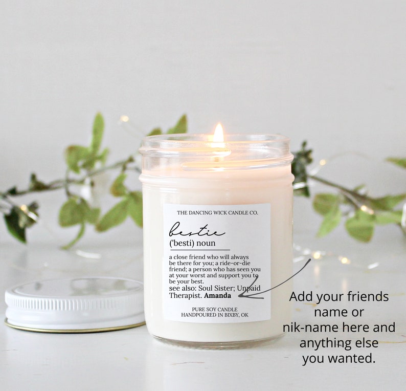 May include: A white candle in a glass jar with a white lid. The label on the jar reads "THE DANCING WICK CANDLE CO. bestie ('besti) noun a close friend who will always be there for you; a ride-or-die friend; a person who has seen you at your worst and support you to be your best. see also: Soul Sister; Unpaid Therapist. Amanda PURE SOY CANDLE HANDPOURED IN BIXBY, OK"