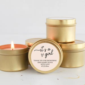 May include: Gold candle tins with a pink and white label that says "it's a girl" with a heart. The label also says "Thank you for showering Erin & Baby Olivia with love 07.25.20xx".