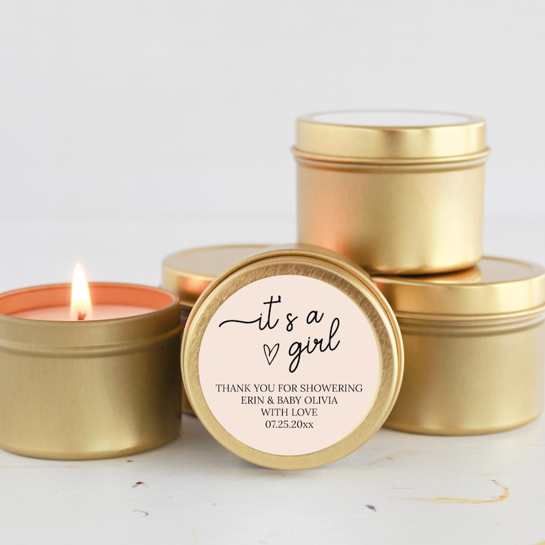 May include: Gold candle tins with a pink and white label that says "it's a girl" with a heart. The label also says "Thank you for showering Erin & Baby Olivia with love 07.25.20xx".