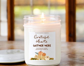 May include: A white soy candle in a clear glass jar with a label that reads "The Dancing Wick Candle Co. Grateful Hearts Gather Here 8 oz Pure Soy Candle Handpoured in Bixby OK". The label features a watercolor illustration of pumpkins and hay bales.