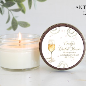 May include: A white candle in a clear glass jar with a gold lid. The lid has a label with a champagne glass and pearls, and the text "Emily's Bridal Shower Thank you for celebrating with us! 08.13.20xx".