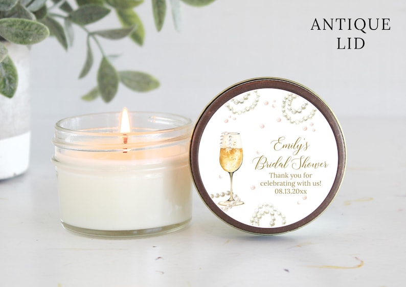 May include: A white candle in a clear glass jar with a gold lid. The lid has a label with a champagne glass and pearls, and the text "Emily's Bridal Shower Thank you for celebrating with us! 08.13.20xx".
