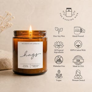 May include: A 9 oz amber glass jar candle with a white label reading "hugs" and "THE DANCING WICK CANDLE CO." A lit wick emits a thin wisp of smoke. To the right, icons and text list features: Pure Soy Wax, Hand Poured, All Natural Ingredients, 100% Cotton Wick, Phthalate Free, Made in USA, Vegan, and Woman Owned.