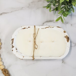 May include: An oval-shaped, distressed white wooden dough bowl candle filled with smooth white wax and three wicks. The bowl is tied with a thin piece of brown twine wrapped around the center. A strand of natural wooden beads lies nearby.
