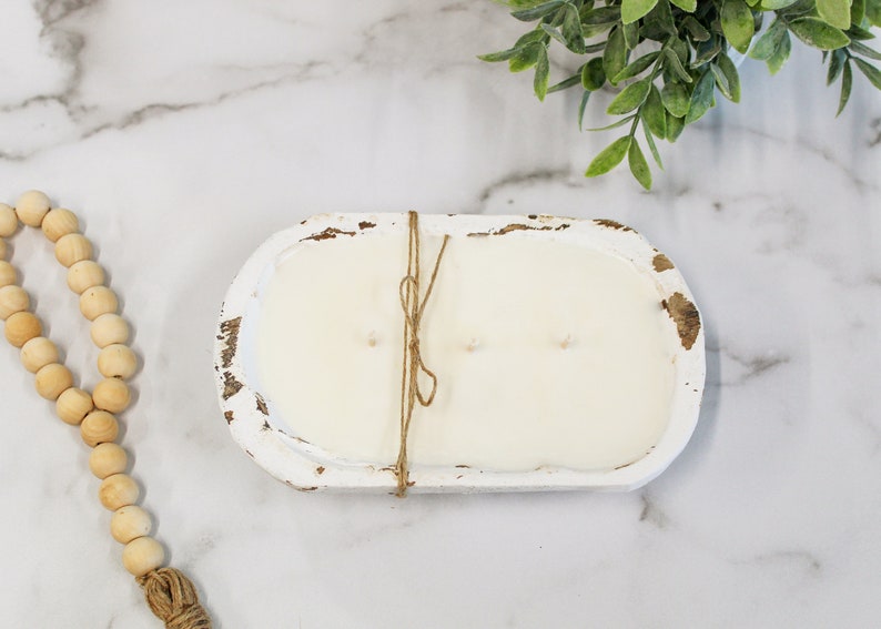 May include: An oval-shaped, distressed white wooden dough bowl candle filled with smooth white wax and three wicks. The bowl is tied with a thin piece of brown twine wrapped around the center. A strand of natural wooden beads lies nearby.