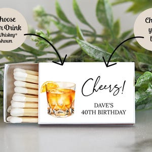 Puede incluir: Una caja de cerillas blanca con un dise&ntilde;o de un vaso de whisky con una rodaja de lim&oacute;n. El texto "Cheers! Dave's 40th Birthday" est&aacute; impreso en la caja de cerillas.