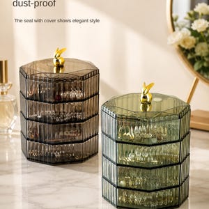 May include: Two multi-tiered, octagonal jewelry organizers made of translucent, ribbed plastic. One container is dark gray and the other is sage green. Each features four rotating storage compartments and a gold-colored rabbit figurine on the lid.