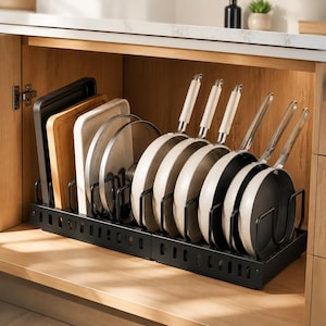 May include: A black metal kitchen organizer rack holding several light-colored frying pans with long handles, circular pot lids, a wooden cutting board, and a dark rectangular baking sheet, all arranged vertically within a wooden cabinet shelf.