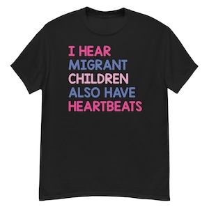 Könnte beinhalten: Ein schwarzes kurzärmeliges T-Shirt mit dem Text I HEAR MIGRANT CHILDREN ALSO HAVE HEARTBEATS, der in abwechselnden Zeilen aus rosa und hellblauen Großbuchstaben mittig auf der Vorderseite aufgedruckt ist.