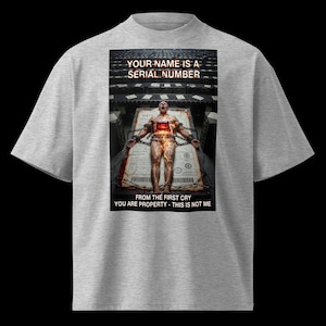 May include: A grey t-shirt with a graphic design depicting a person chained to a document. The text on the shirt reads "YOUR NAME IS A SERIAL NUMBER" and "FROM THE FIRST CRY YOU ARE PROPERTY - THIS IS NOT ME."