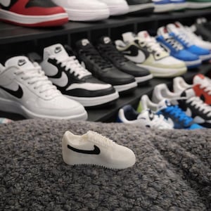 Può includere: Una sneaker bianca in miniatura con un logo nero a virgola è appoggiata su una superficie grigia e strutturata. Gli scaffali sullo sfondo mostrano una varietà di sneaker in diversi colori, tra cui rosso, nero, bianco e blu.