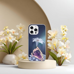 May include: A smartphone case featuring a graphic illustration of a hand holding a single white lily against a dark blue background. The case has a cutout for a triple-lens camera system and is displayed on a cylindrical stand.