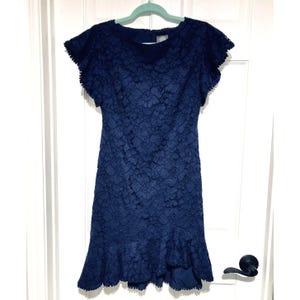 May include: A navy blue lace dress with short, ruffled sleeves and a ruffled hem. The dress is hanging on a light green hanger against a white door. The dress has a round neckline and a fitted silhouette.