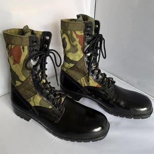 May include: A pair of high-top combat boots featuring black leather lower sections and camouflage patterned fabric uppers in shades of green, brown, and tan. The boots include black metal eyelets, black laces, and a reinforced black rubber sole.